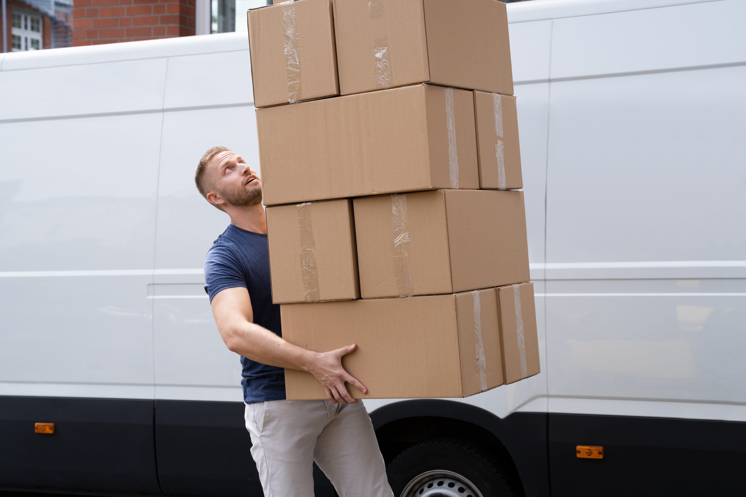 A man struggles to balance a tall stack of five cardboard boxes while walking beside a white moving van.