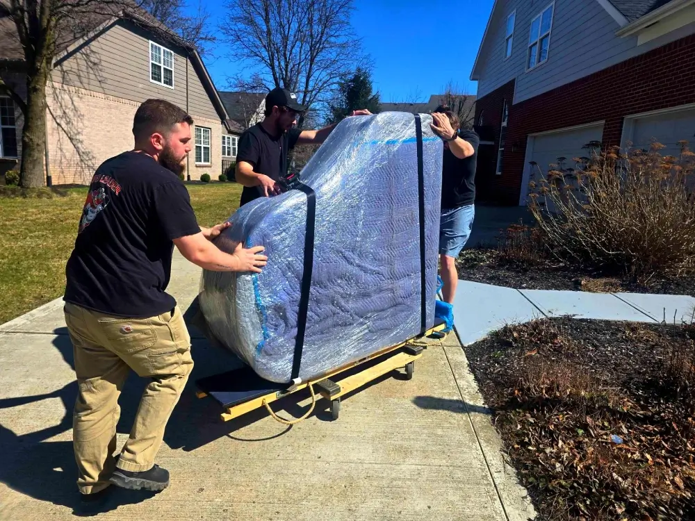 Moving crew guiding a securely wrapped piano on a moving dolly outside a home.