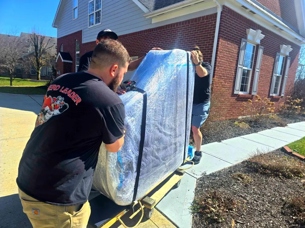 Three movers carefully transporting a wrapped and protected piano outside a residential home.